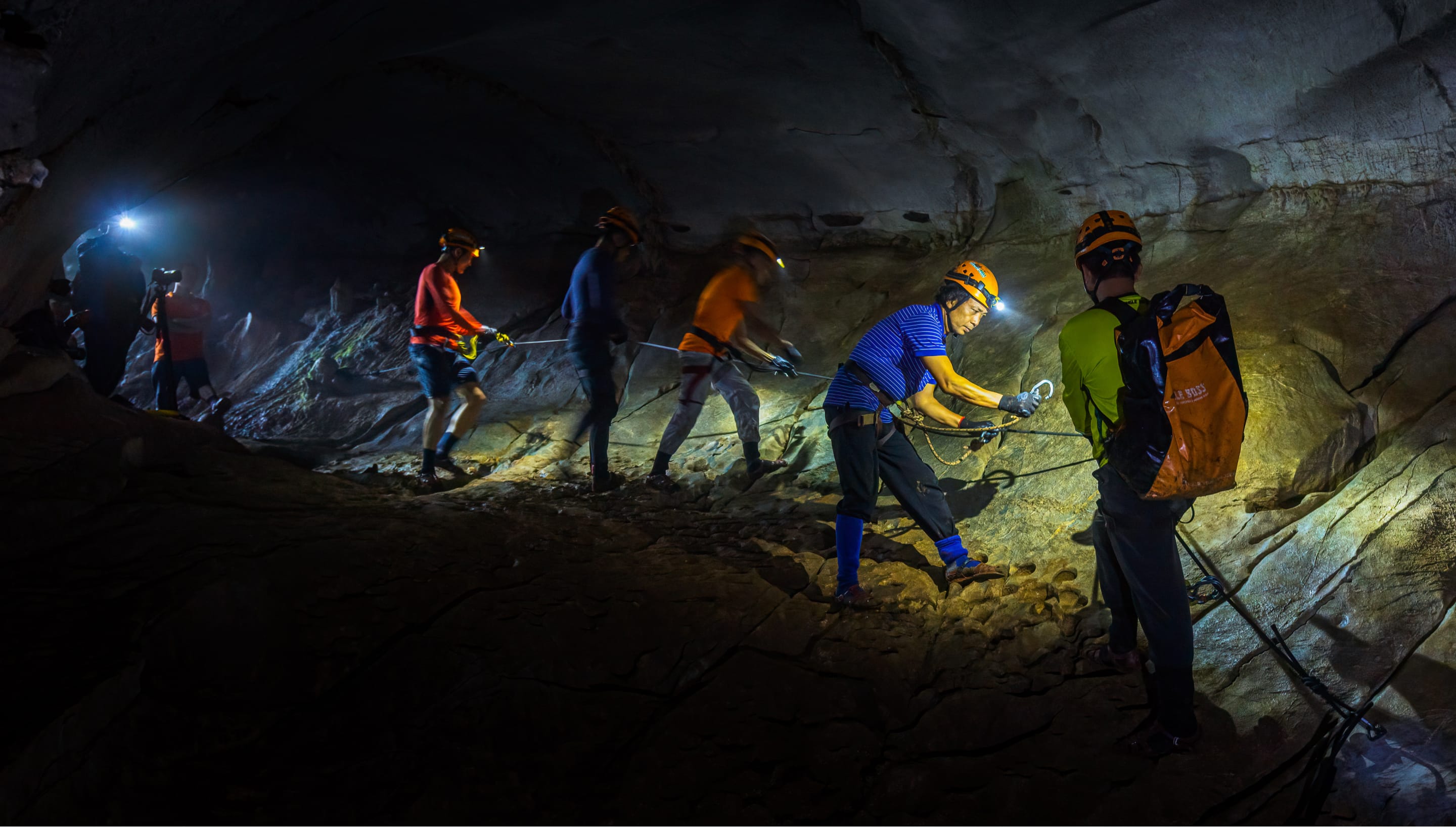 Trekkers in helmets and head torches moving roped through a dark cave passage