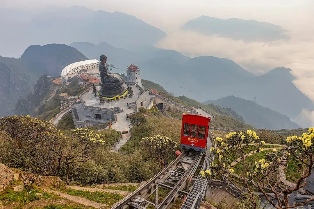 The red Fansipan summit funicular train climbing the tracks past the giant bronze Great Amitabha Buddha statue in Sapa.