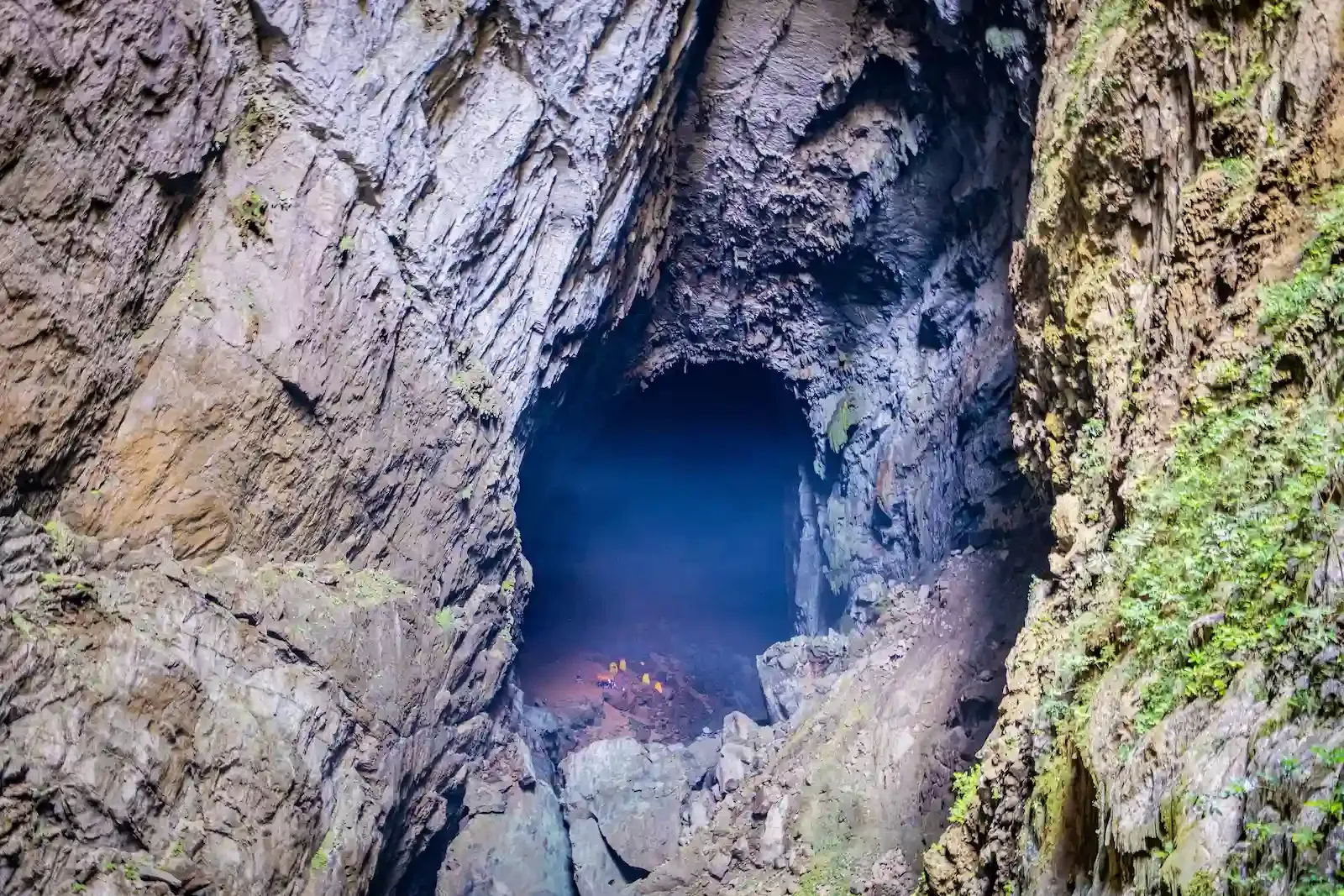High viewpoint looking down into the dramatic entrance shaft of Son Doong Cave with tiny orange tents glowing on the rocky floor far below in Vietnam