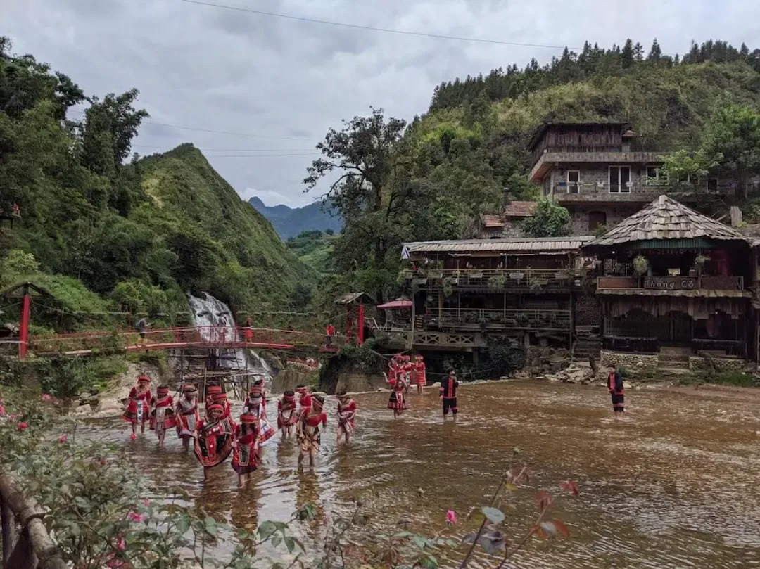Village de Cat Cat près de Sapa au Vietnam, habitants Hmong en tenues traditionnelles rouges dans la rivière devant cascade, pont et maisons en bois de montagne