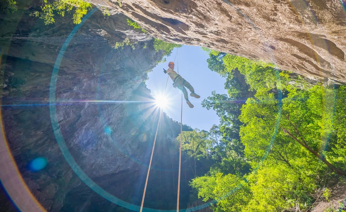 A climber abseiling on a rope into a jungle sinkhole with the sun behind the rim