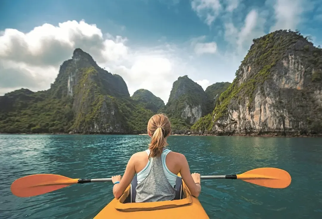 Woman Paddling Kayak Through Pristine Turquoise Waters of Bai Tu Long Bay With Dramatic Limestone Mountains in Background - Peaceful Kayaking Adventure