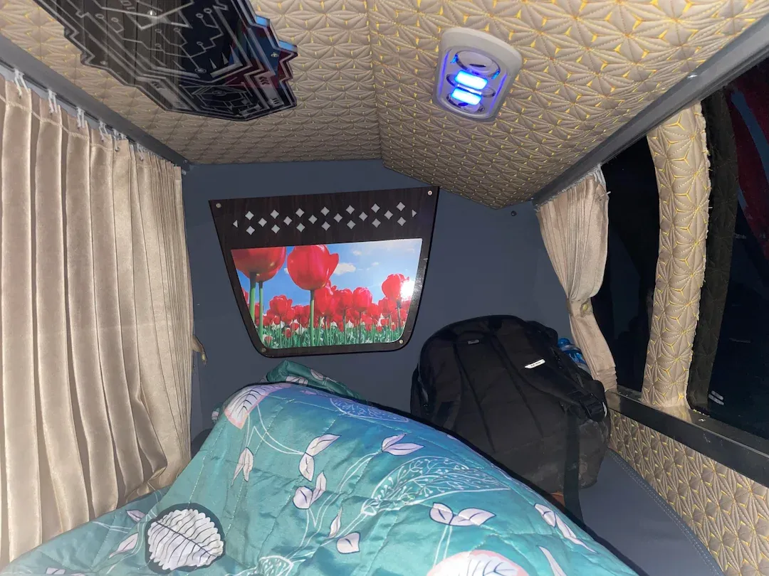 Comfortable interior view of a Vietnam sleeper bus cabin with a turquoise and white patterned bed, cream-colored curtains, and decorative textured ceiling with blue accent lighting