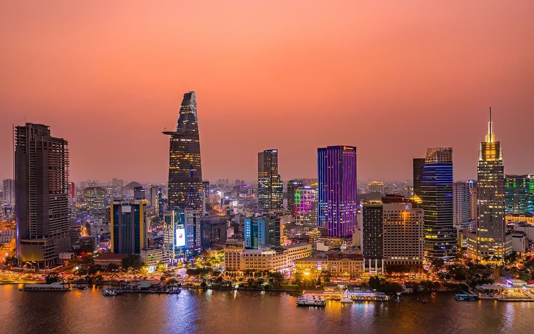 a Vibrant Cityscape of Ho Chi Minh City at Dusk, Featuring Illuminated Skyscrapers Reflecting on the Saigon River Under a Warm, Orange Sky