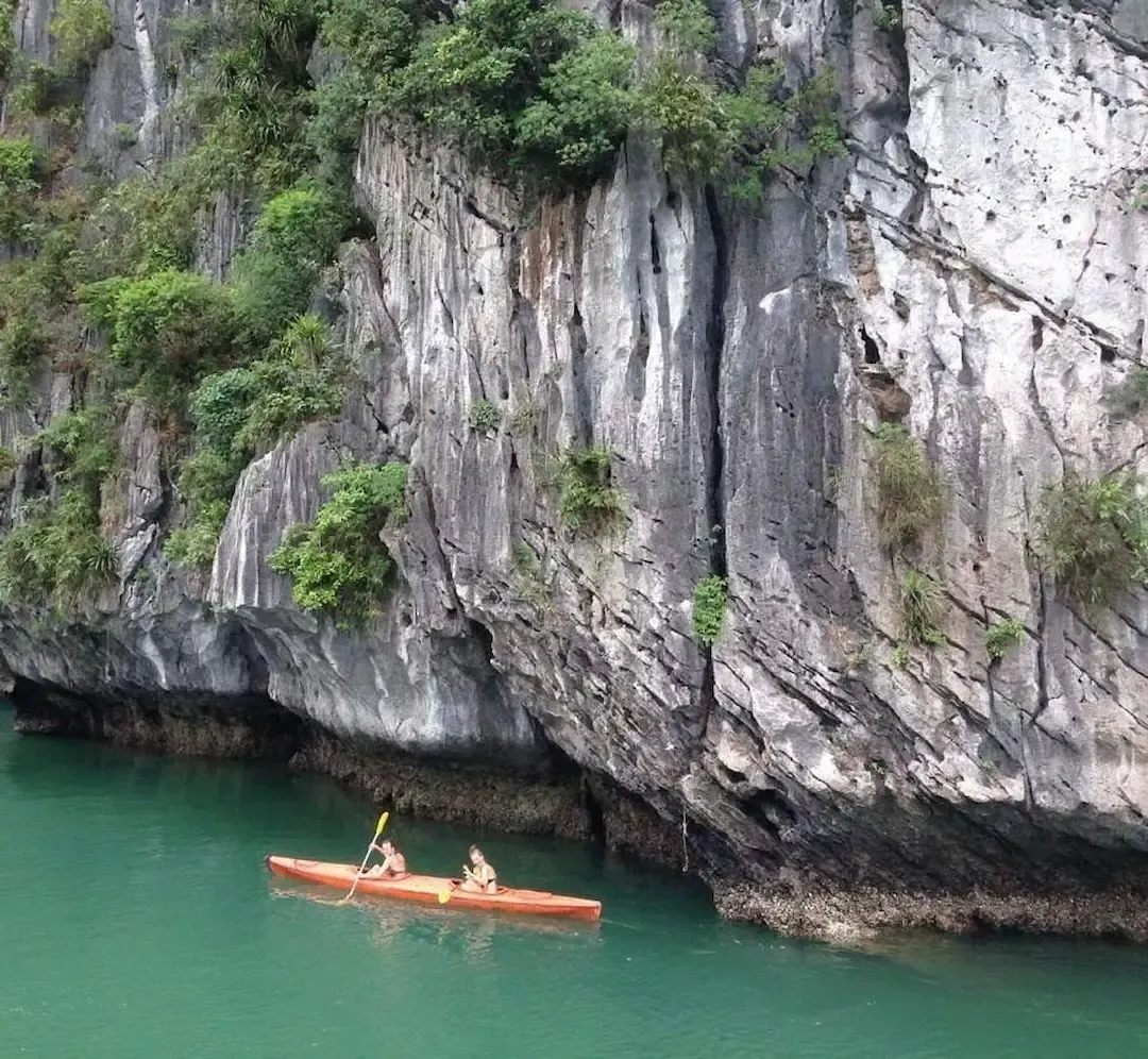 Orange Kayak Gliding Beside Steep Limestone Cliff Covered in Greenery on Emerald Waters of Lan Ha Bay