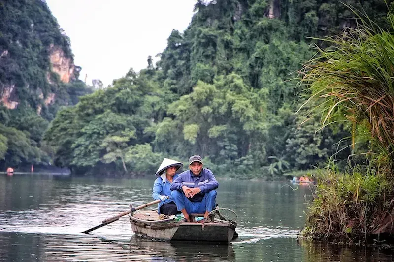 Local rowers using traditional bamboo poles to navigate wooden boat through Trang An river