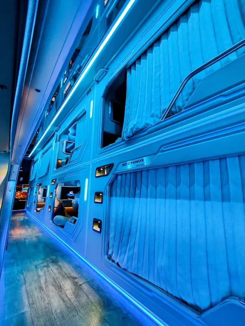 Modern Vietnam sleeper bus interior with blue neon ambient lighting, corrugated metal walls, individual bunk beds with side reading lights, and premium cabin design