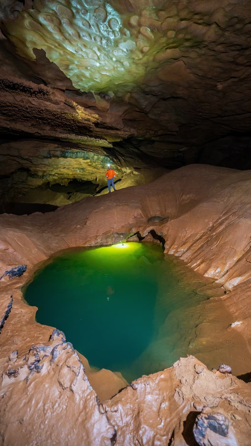 a Person Stands on a Rocky Ledge Inside a Vast Cave, Overlooking a Vibrant Green Underground Pool Illuminated by Artificial Light in Hung Thoong Cave System