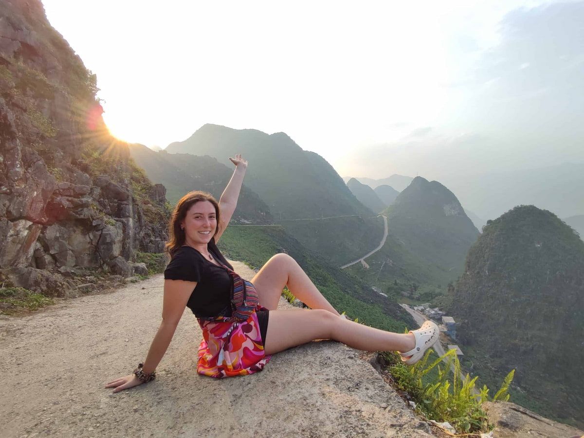 Person Sitting on a Mountain Edge at Sunset in Ha Giang, Vietnam, Smiling and Pointing at Distant Peaks