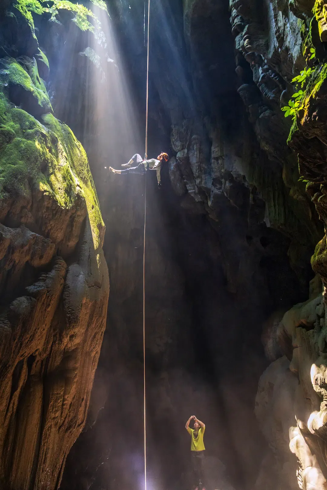 Explorer crossing a slackline above Kong Collapse sinkhole during the Tiger Cave Series expedition in Phong Nha-Ke Bang National Park, with sunlight streaming through the opening above.