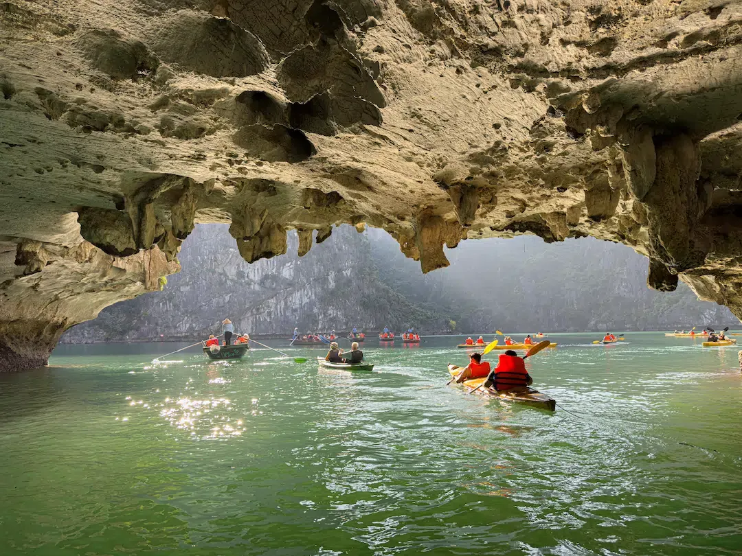 Tourists paddling kayaks through a spectacular limestone cave archway over emerald water in Lan Ha Bay.