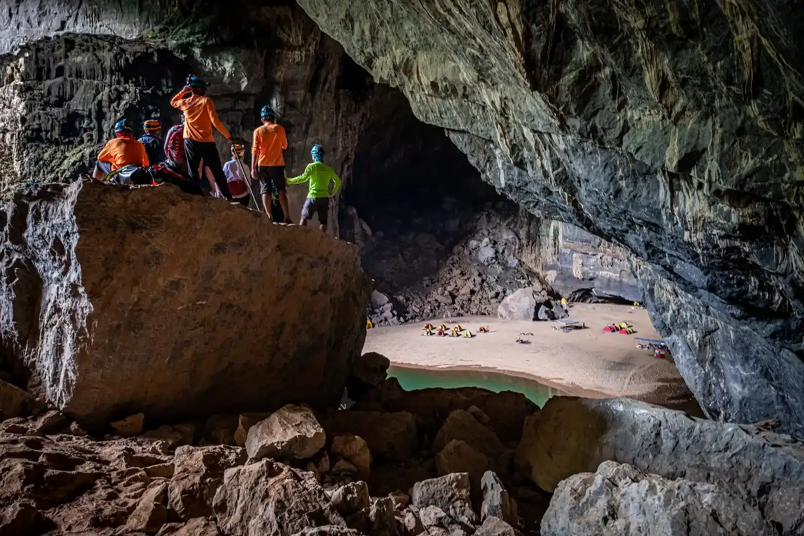 Adventure group standing on boulders overlooking an underground beach campsite and emerald pool inside Son Doong Cave on a guided expedition in Phong Nha Ke Bang Vietnam.