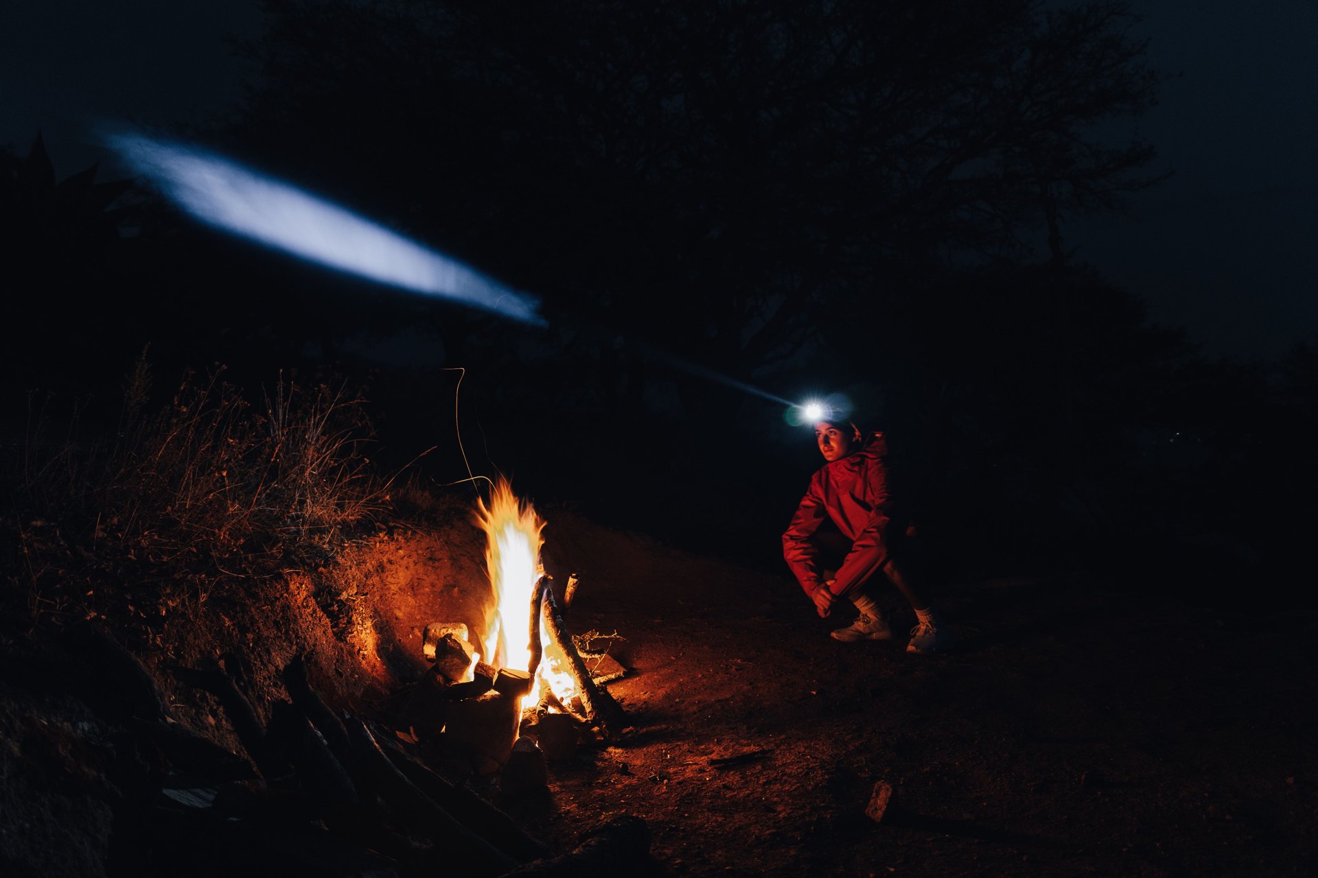 Person wearing a head torch crouched beside a campfire at night