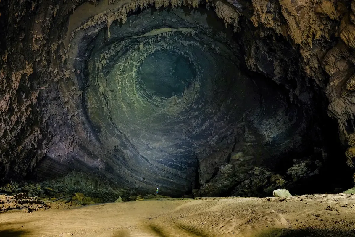 Tiny explorer on the sandy floor of a vast underground chamber in the Tu Lan cave system, Phong Nha, beneath a dramatic circular ceiling dome. 