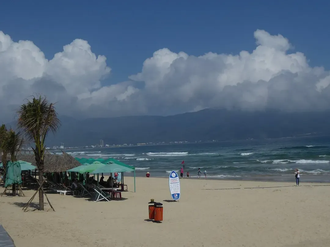 Surfer riding waves at My An surfing beach (Danang) with sandy shoreline, beach vendors with straw umbrellas, and crowds of tourists during peak season