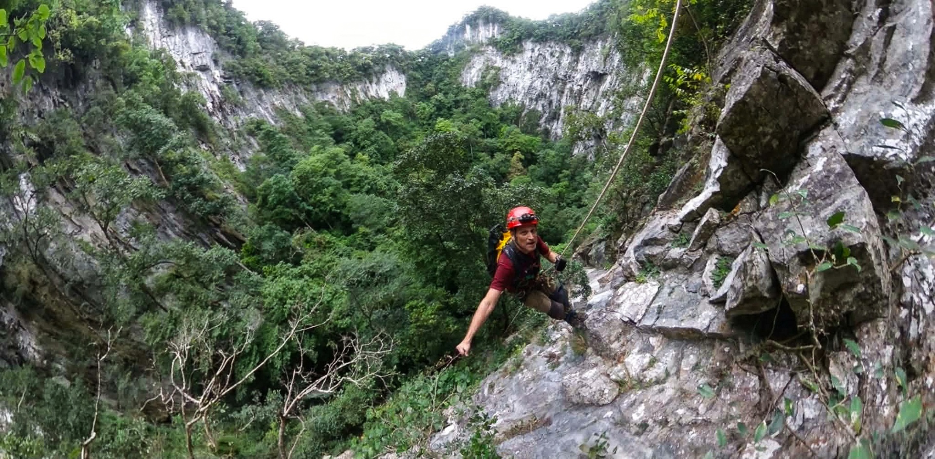 Helmeted caver on a steep rocky slope above the forested sinkhole floor