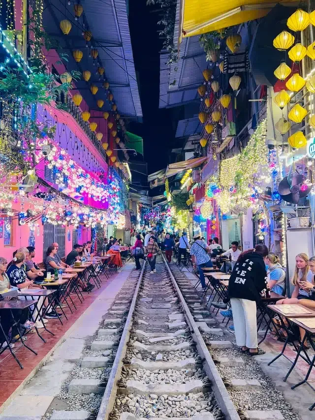 Tourists sitting at outdoor cafe tables along the narrow Hanoi Train Street tracks illuminated by colorful purple and pink neon lights at night.