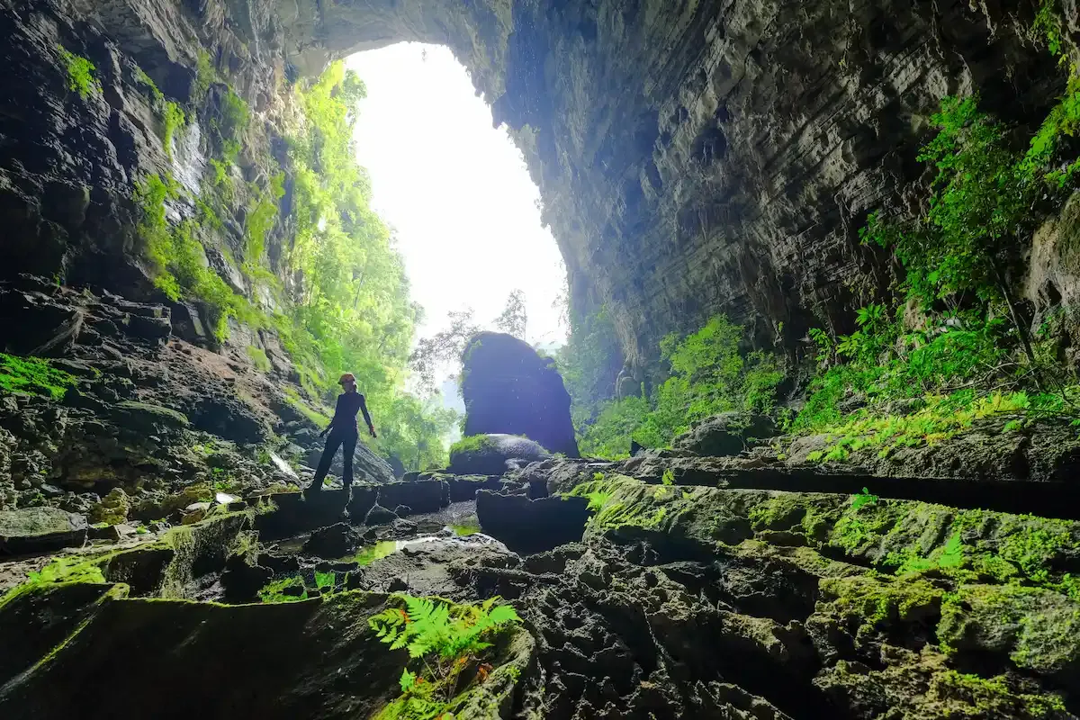 Traveler walking across wet rocky pools beneath the towering skylight of Tu Lan cave in Phong Nha, with bright green jungle spilling inside.