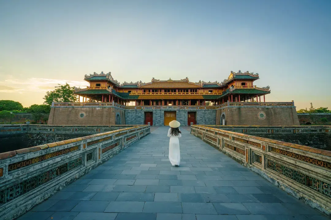 Woman in white ao dai and conical hat walking toward Meridian Gate Ngo Mon at Hue Imperial City UNESCO World Heritage Site Vietnam at sunset