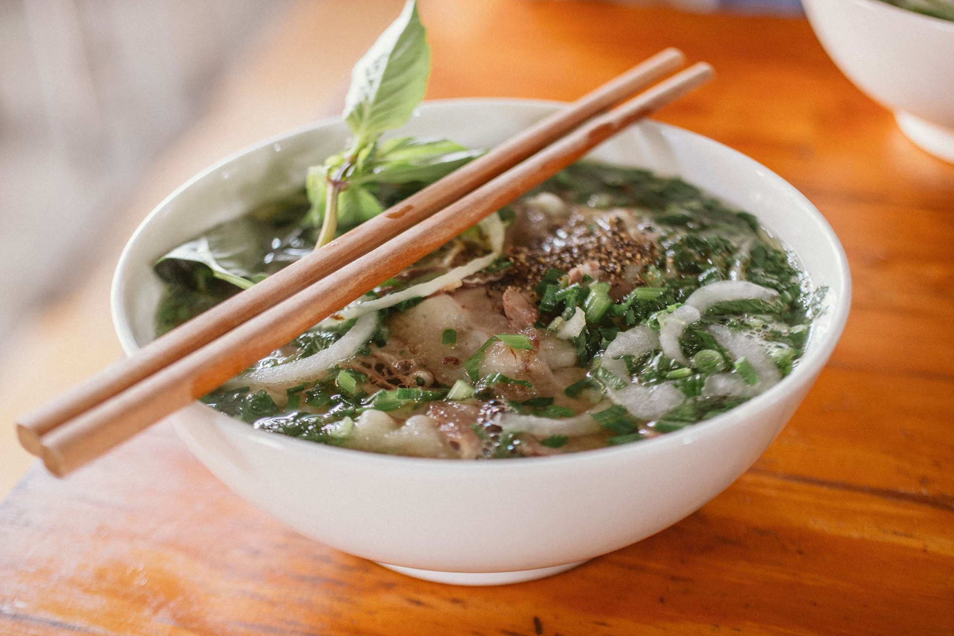 Bowl of beef pho with basil, onion and chopsticks on a wooden table