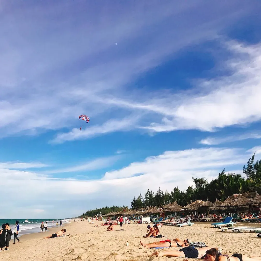 Non Nuoc Beach Da Nang offering parasailing activities with tourist paragliding over blue sea, sandy beach, and coastal mountains in background
