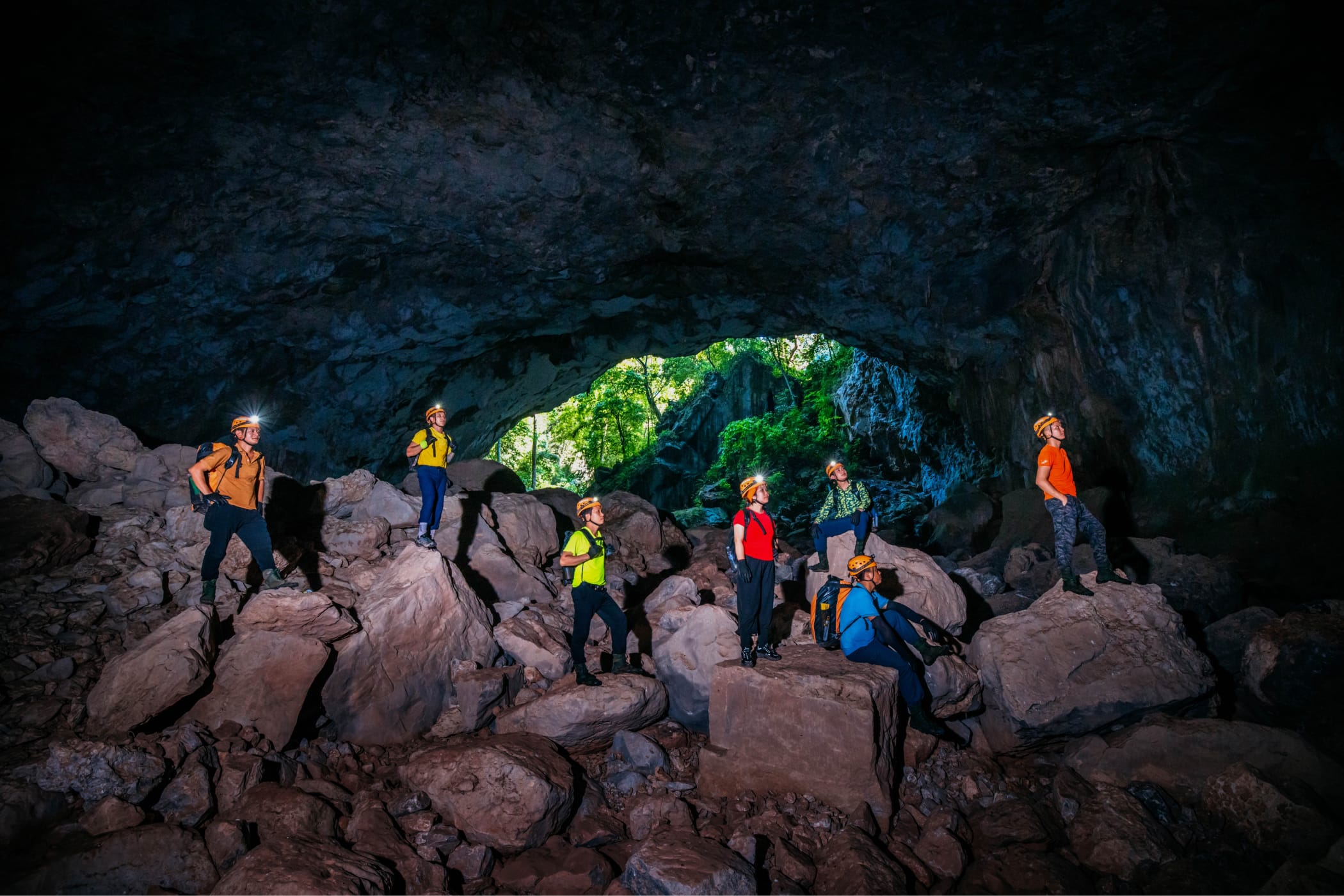 Expedition group resting on boulders at a cave entrance with daypacks