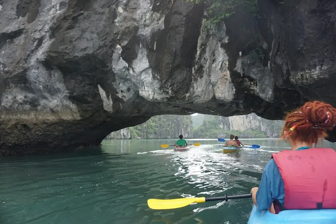 Kayakers Paddling Through Low Limestone Cave Entrance on Calm Emerald Water in Lan Ha Bay