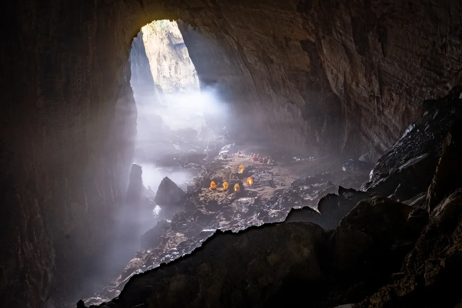 Misty morning inside Son Doong Cave with glowing expedition tents pitched on rocky ground beneath a towering entrance arch in Phong Nha Ke Bang National Park Vietnam