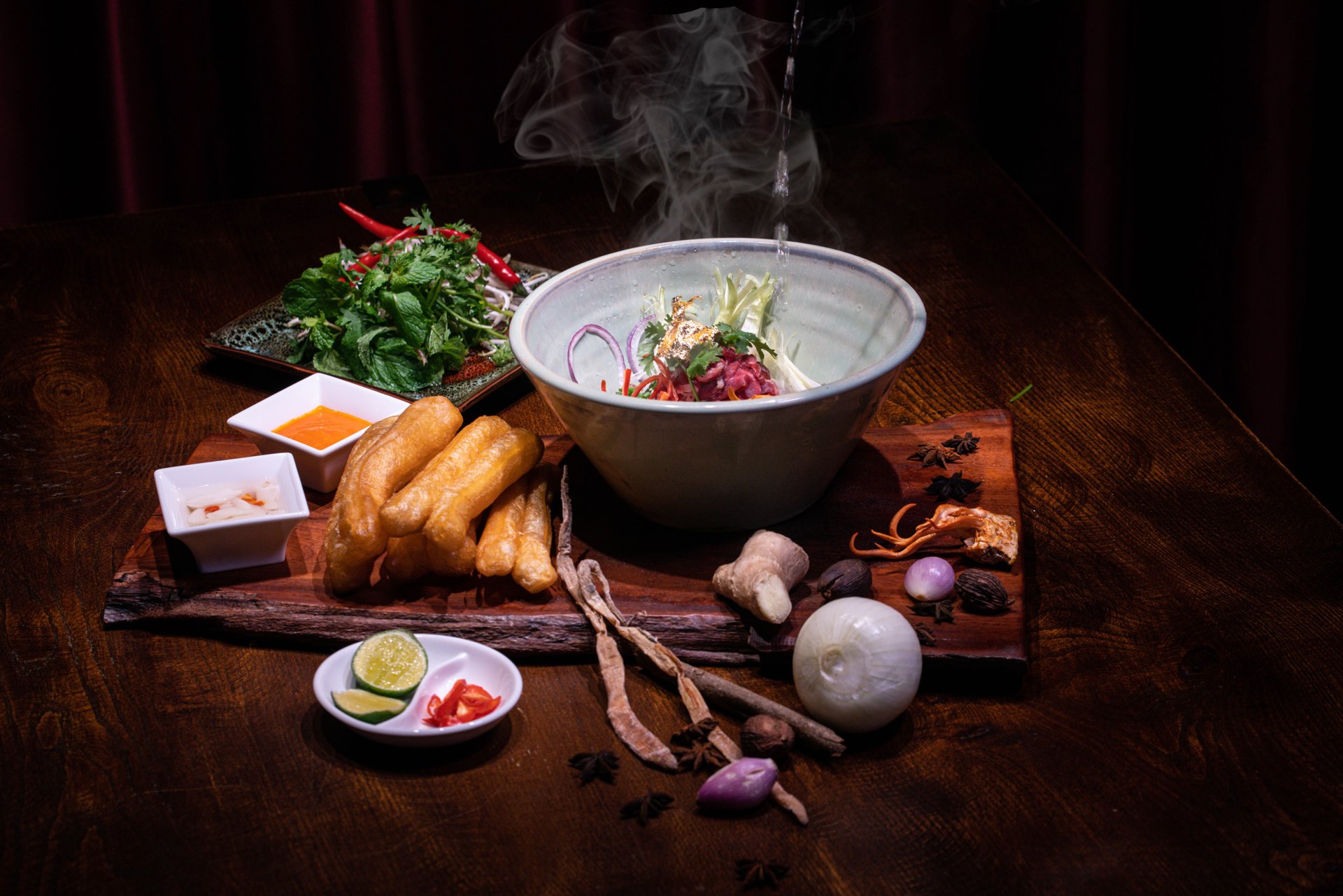Hot liquid poured over beef in a pho bowl beside spices and herbs