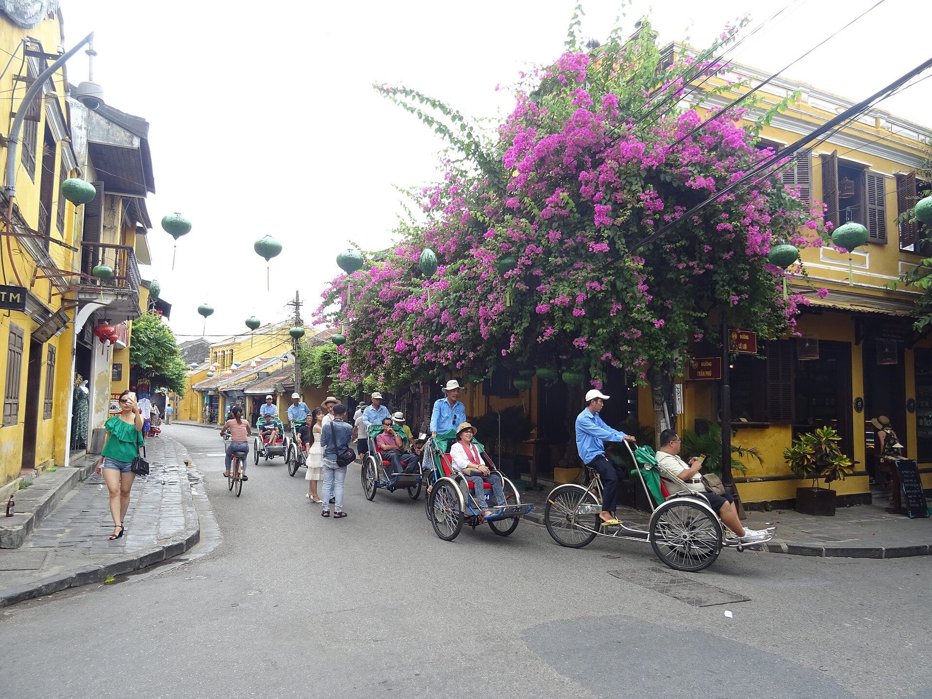 Cyclos and walkers under bougainvillea on a street of yellow houses in Hoi An old town