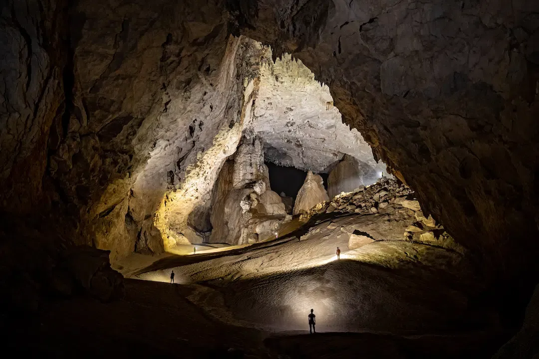 Massive underground cavern chamber view showing tourists with headlamps standing on a sloped sandy floor inside Son Doong Cave