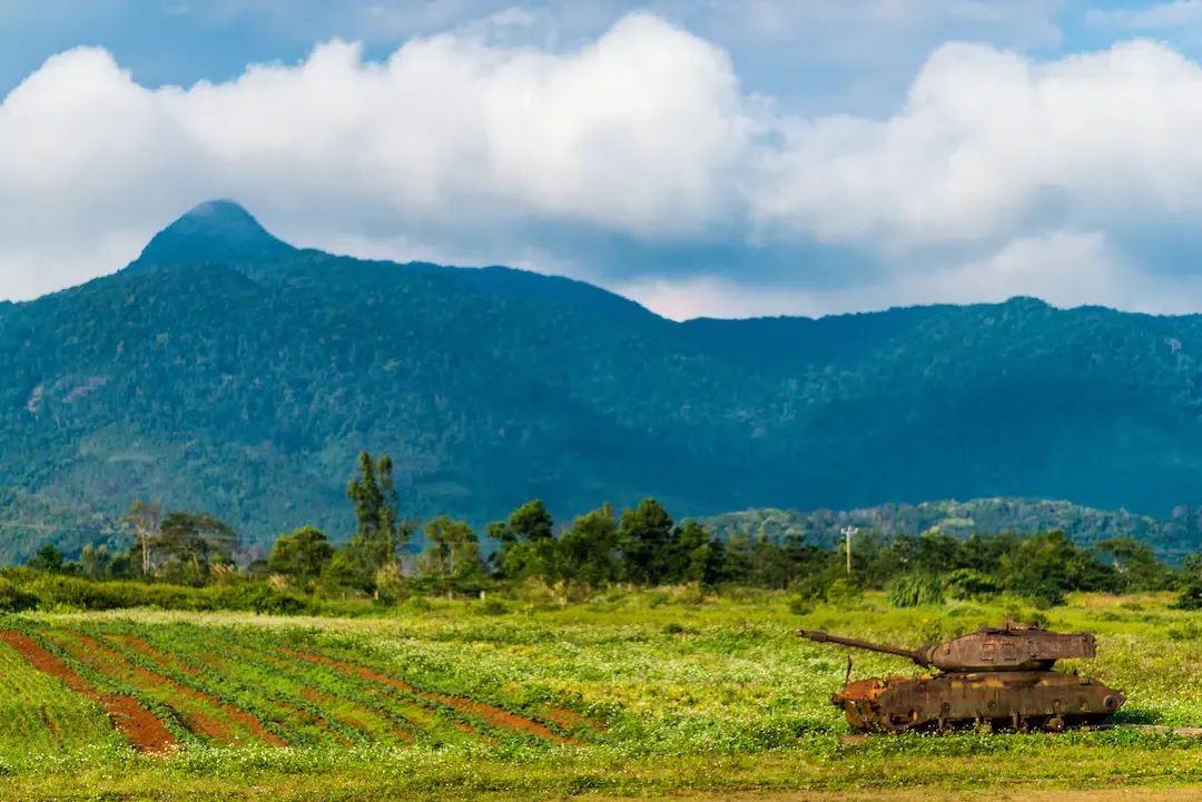 Rusted tank hull in a green field with the misty mountains of Khe Sanh in the background.