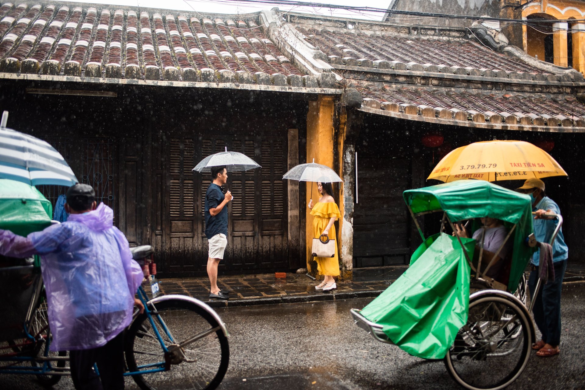 Cyclo drivers in rain ponchos and visitors with umbrellas beside an old tiled house in Hoi An