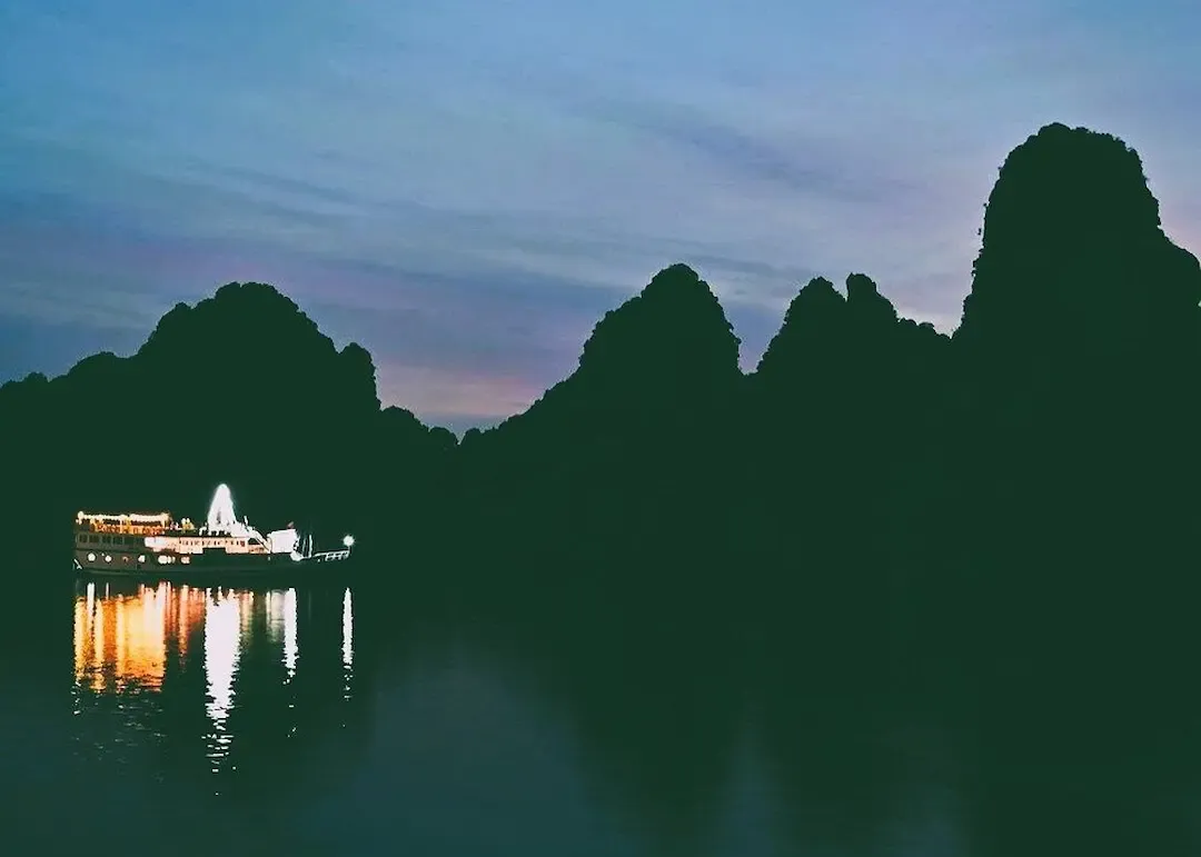 Luxury Cruise Ship Anchored at Bai Tu Long Bay During Twilight Hour With Warm Ship Lighting, Limestone Islands in Background, and Calm Peaceful Waters