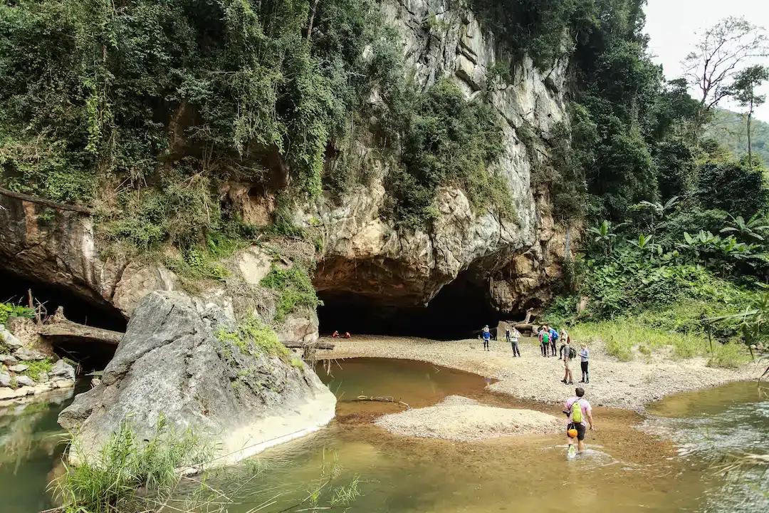 Adventure travelers trekking through a river toward the massive limestone entrance of Hang En Cave in Phong Nha-Ke Bang National Park, Vietnam.
