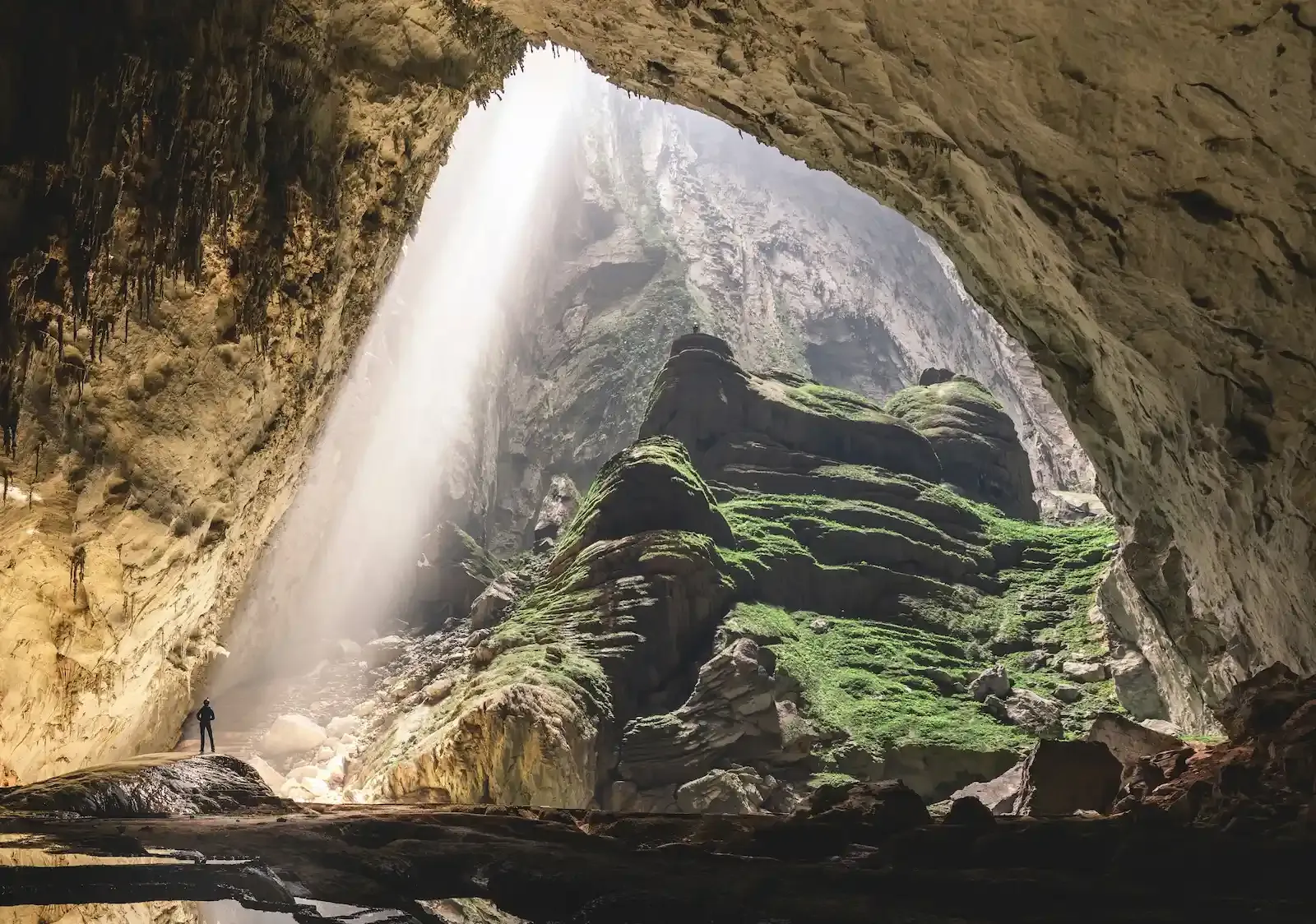 Powerful sunbeam cutting through the ceiling of Son Doong Cave to spotlight layered green terraces and a small caver exploring the world’s largest cave in Vietnam.