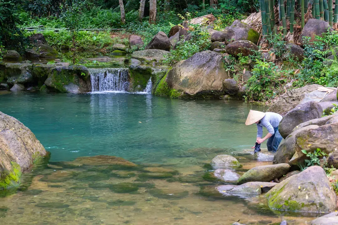 A local woman in a traditional conical hat washing clothes by a small waterfall and stream in Pu Luong Nature Reserve