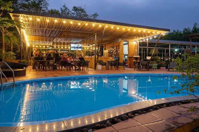 Outdoor Restaurant With a Lit up Pergola Next to a Swimming Pool at Dusk, Featuring Guests Dining and Relaxing.