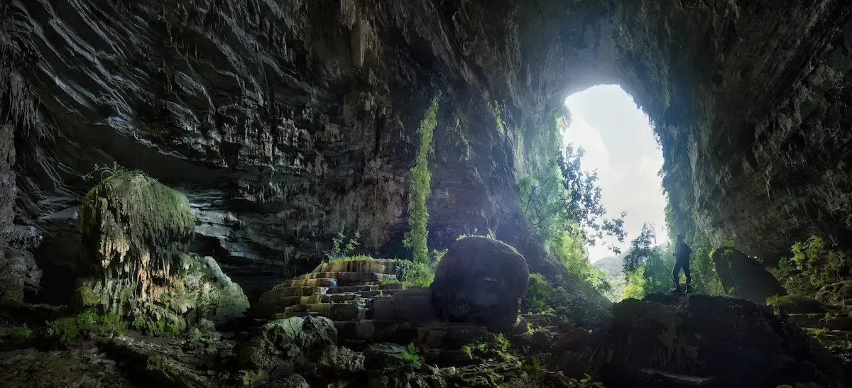 Adventurer standing in the giant entrance chamber of Tu Lan cave in Phong Nha, Vietnam, framed by mossy rocks and jungle daylight.