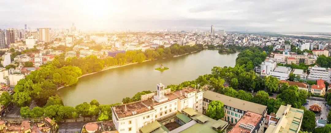 High angle panoramic aerial view of the long green basin of Hoan Kiem Lake surrounded by dense city buildings