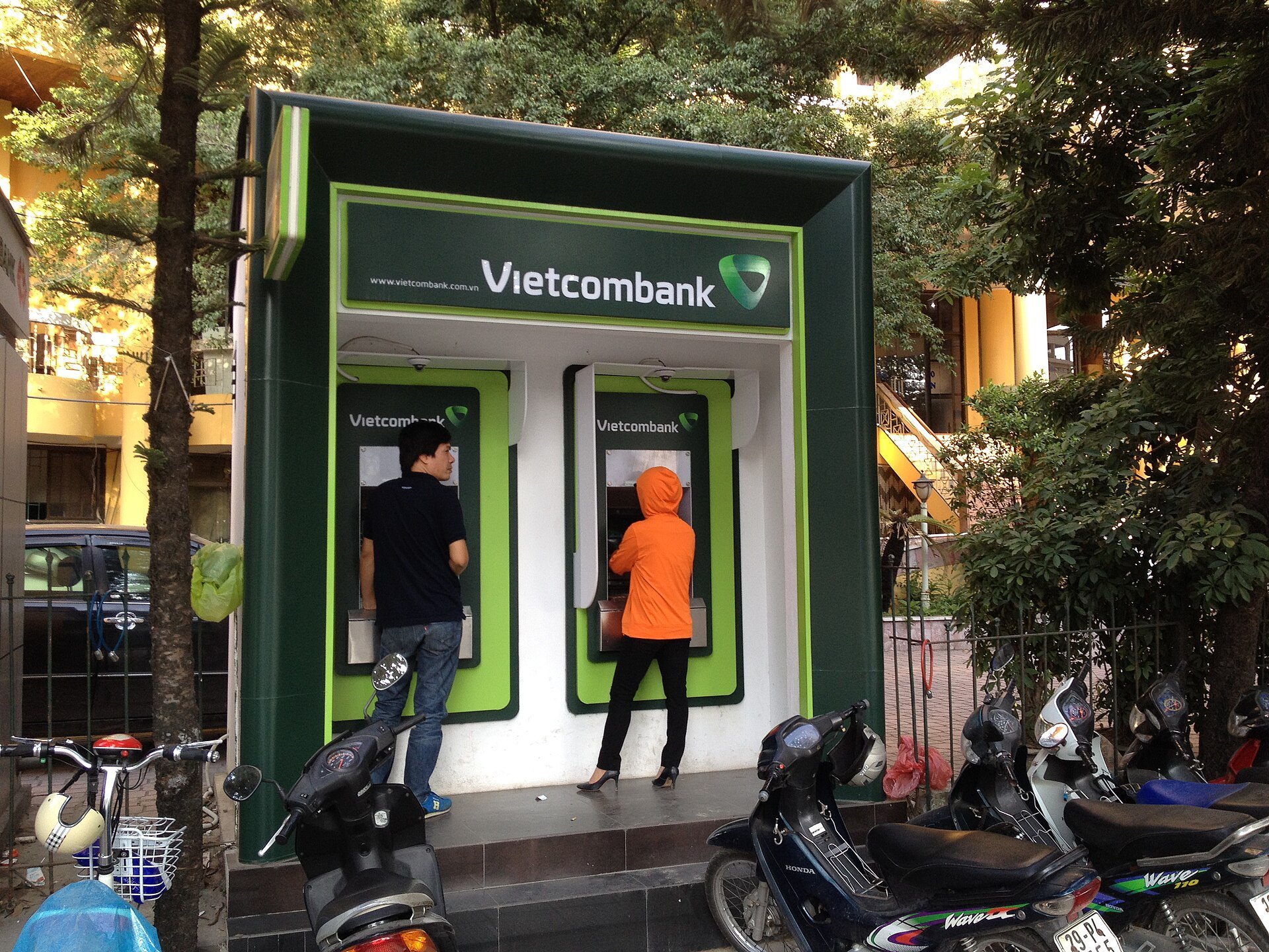 Two people using Vietcombank ATMs in a green booth on a street in Hanoi, with motorbikes parked in front