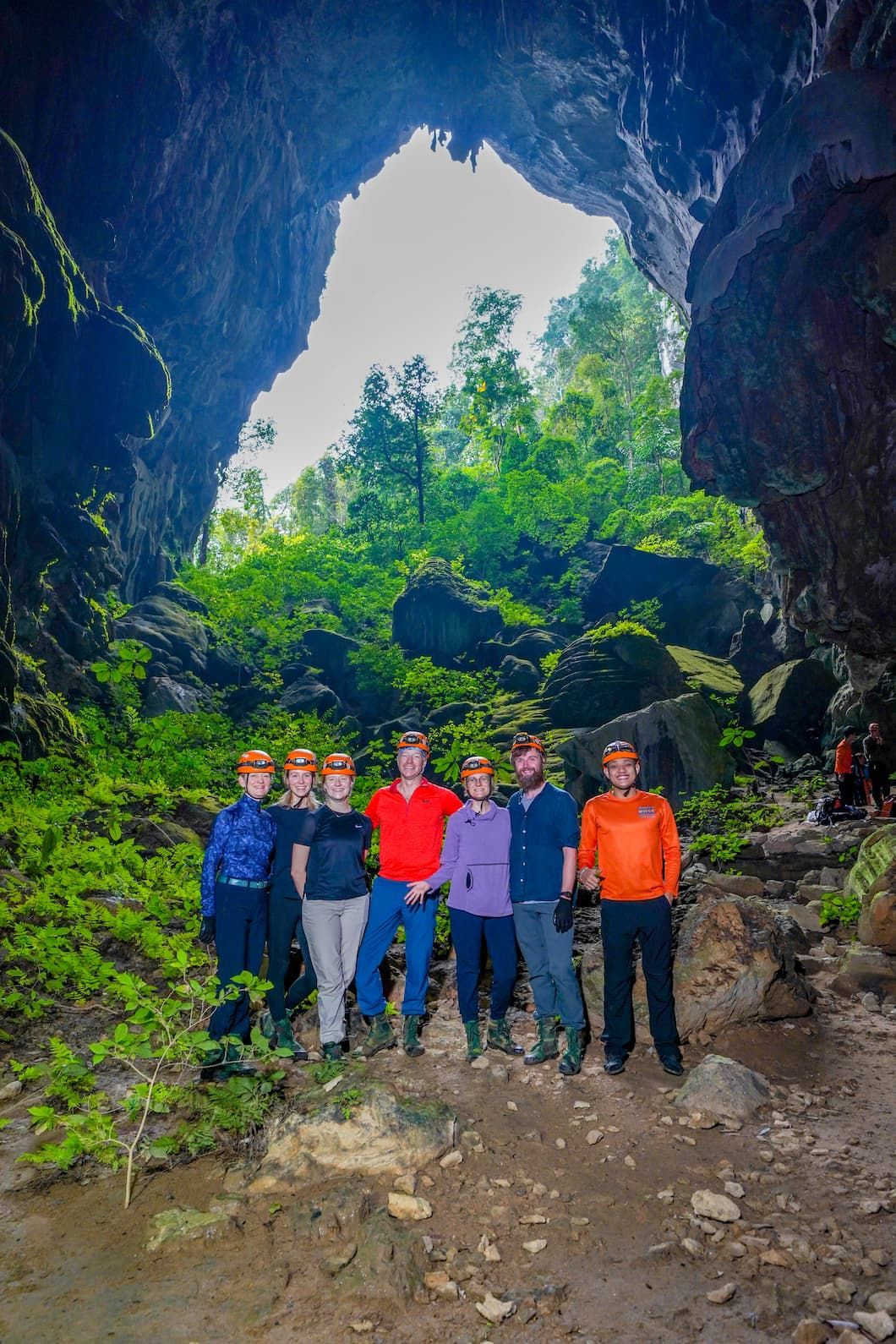 a Group of Seven Travelers Wearing Helmets and Outdoor Gear, Posing Inside a Large, Open Cave Entrance With Lush Green Vegetation and Rocky Terrain in the Background.