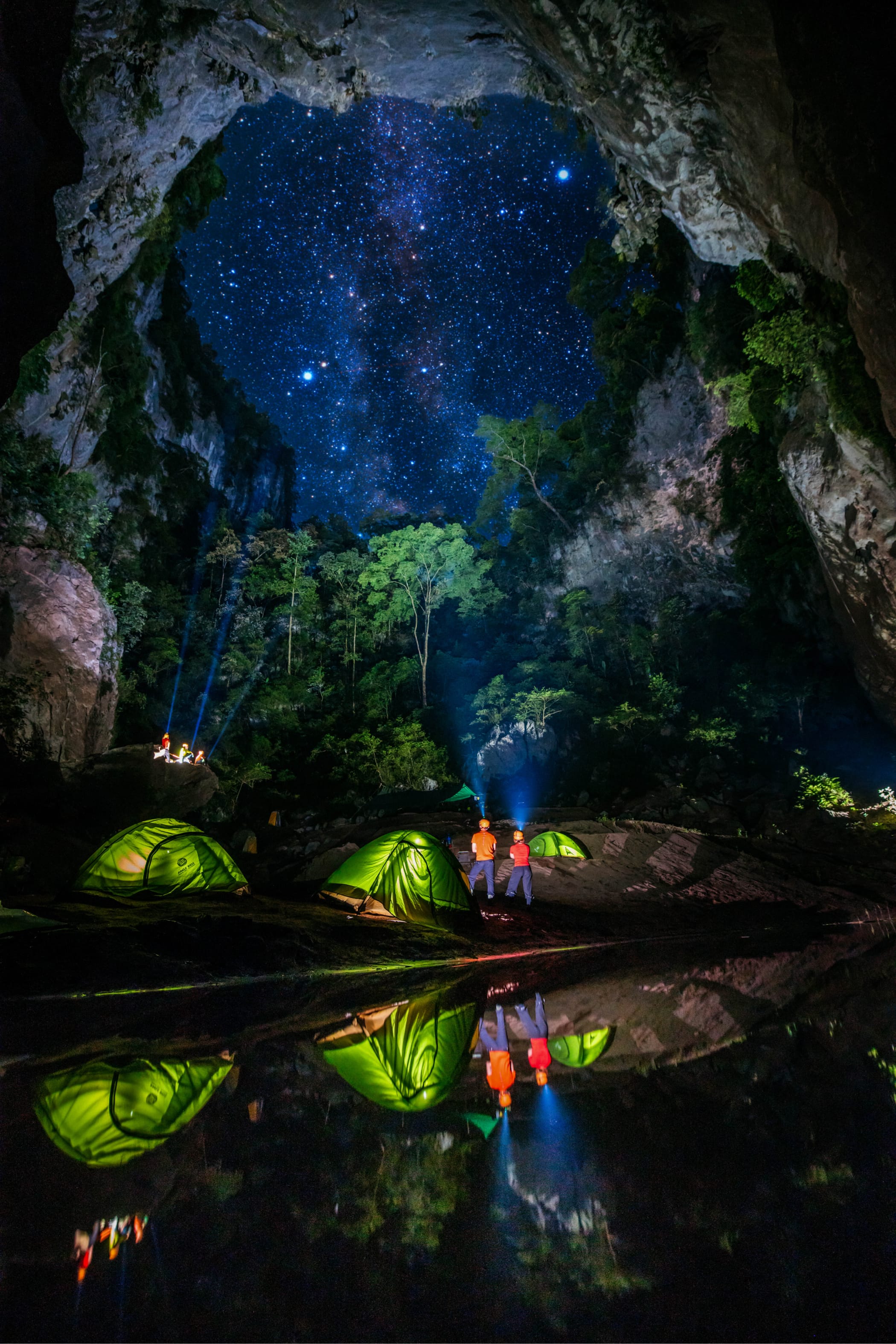 Expedition tents at a cave mouth under a star field, figures by the water