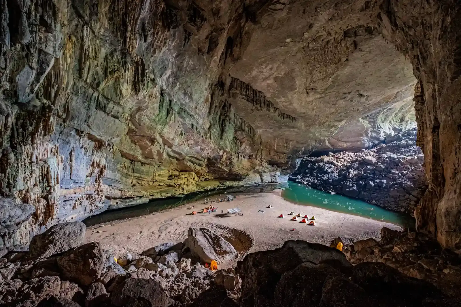 A high-angle view of a vast limestone cave chamber in Hang En, Vietnam, featuring a sandy beach with several colorful camping tents next to a turquoise pool.