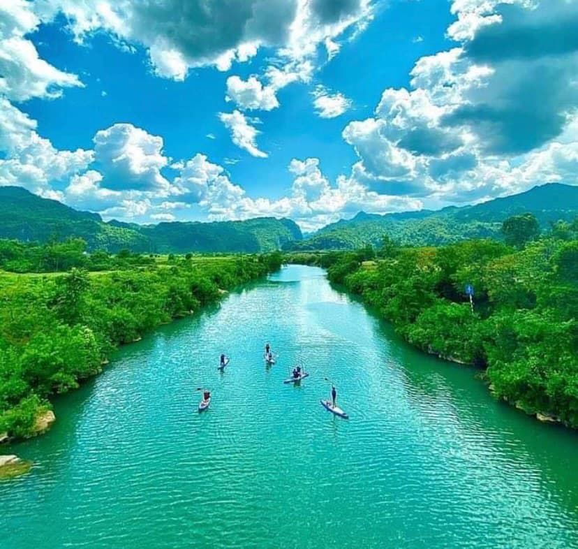 People Paddleboarding on a Serene Chay River Surrounded by Lush Greenery and Mountains Under a Bright Blue Sky With Fluffy Clouds.