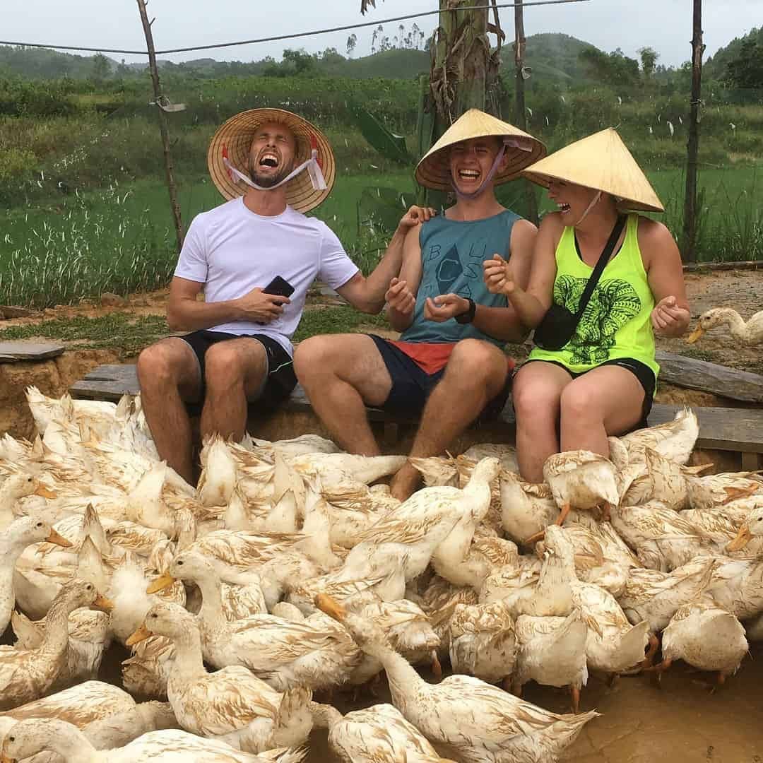 Three People Laughing While Ducks Surround Their Feet at the Duck Stop in Phong Nha