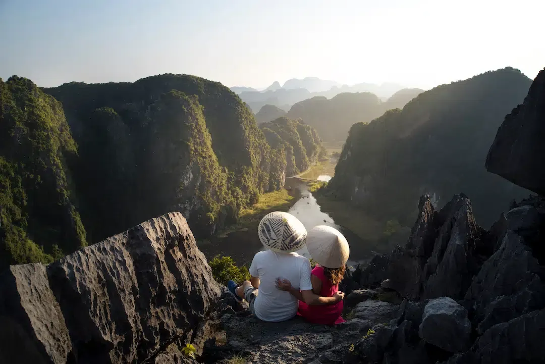 Couple wearing traditional Vietnamese conical hats sitting on Mua Cave summit viewpoint overlooking river valley and endless karst limestone mountains in Ninh Binh Vietnam