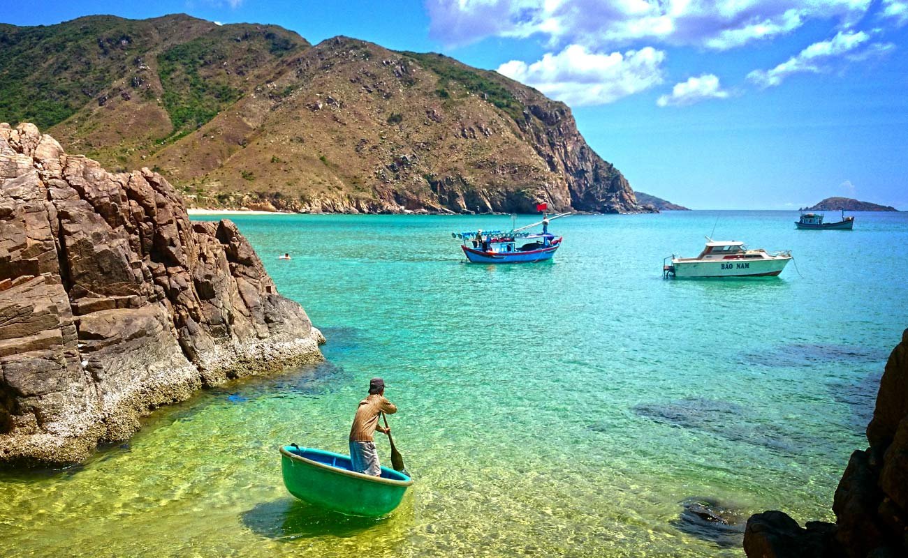 A fisherman paddling a round basket boat in clear turquoise water at Ky Co, below a brown headland