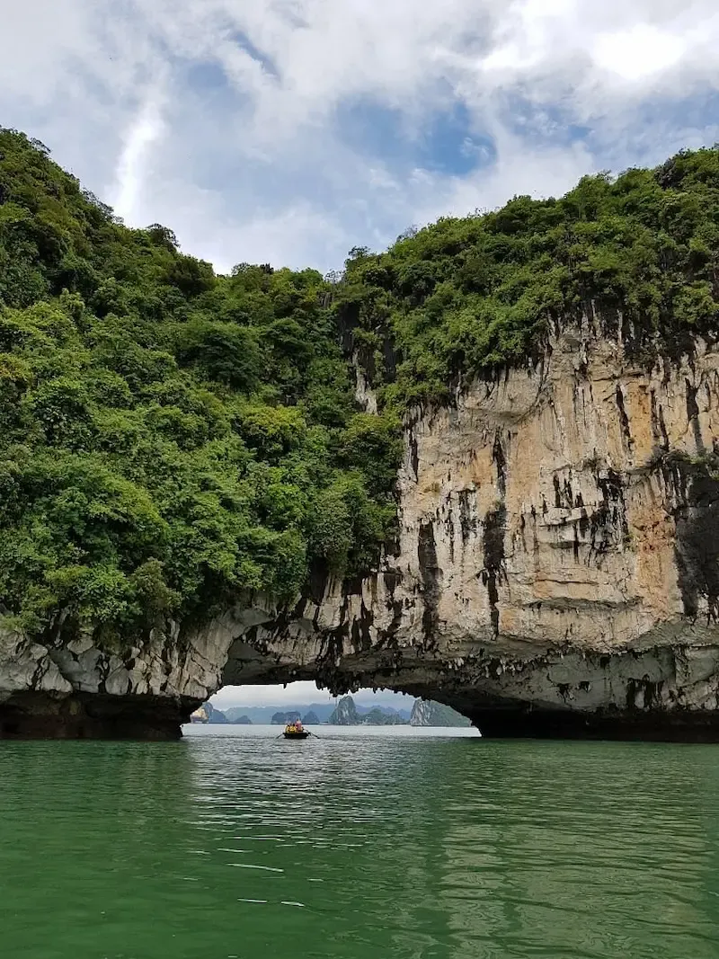 Natural Limestone Arch Tunnel at Bai Tu Long Bay With Emerald Green Water, Jungle Vegetation, and Traditional Boat Passing Through   Prime Kayaking Destination